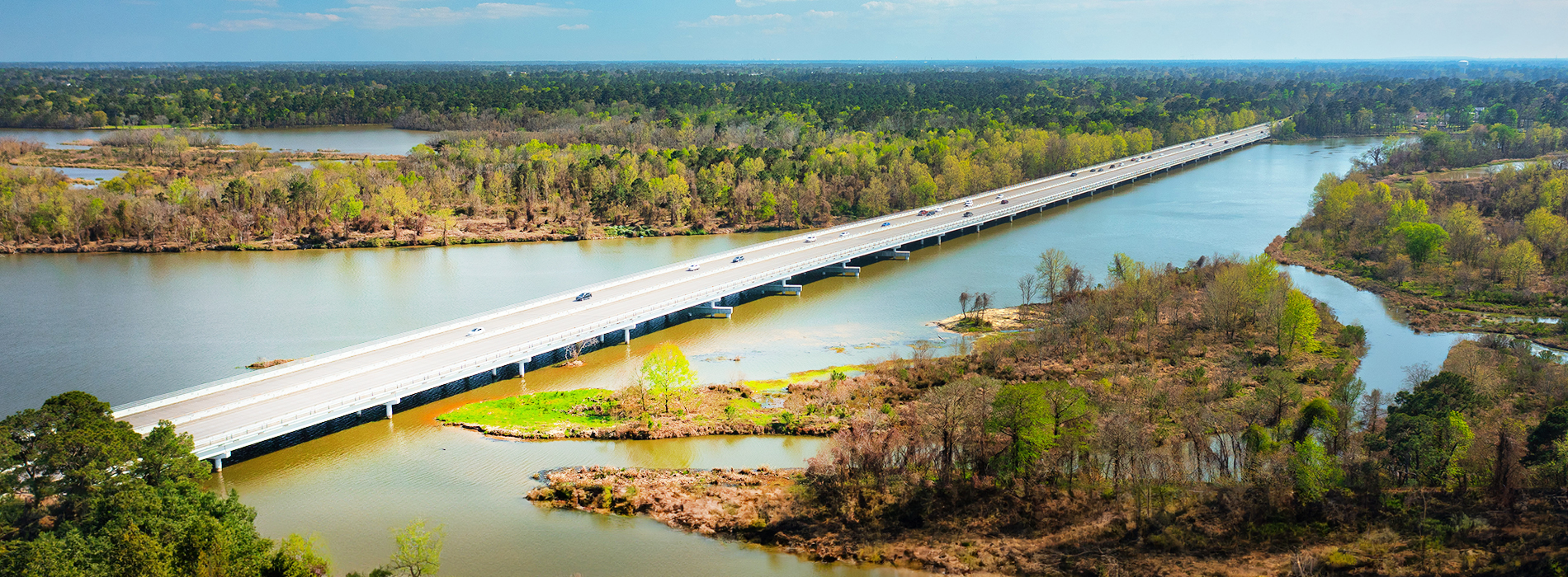 The image depicts a long bridge stretching across a river, with a clear view of the surrounding natural landscape featuring trees and water.