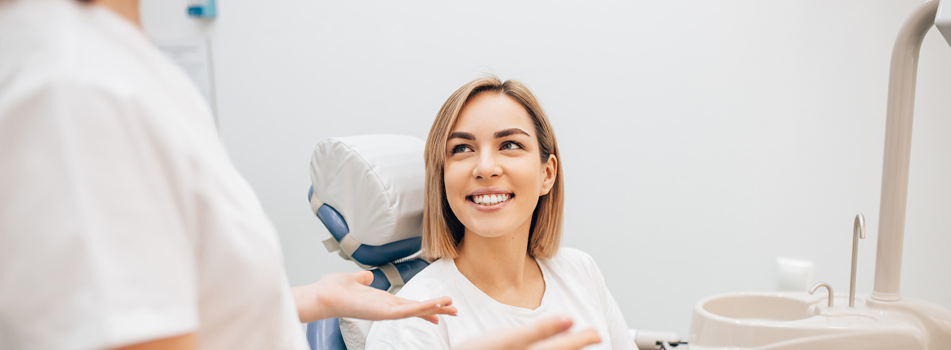 The image shows a dental office setting with a person sitting in a dental chair, receiving treatment, while another individual, possibly a dentist or dental assistant, stands nearby, smiling at the camera.