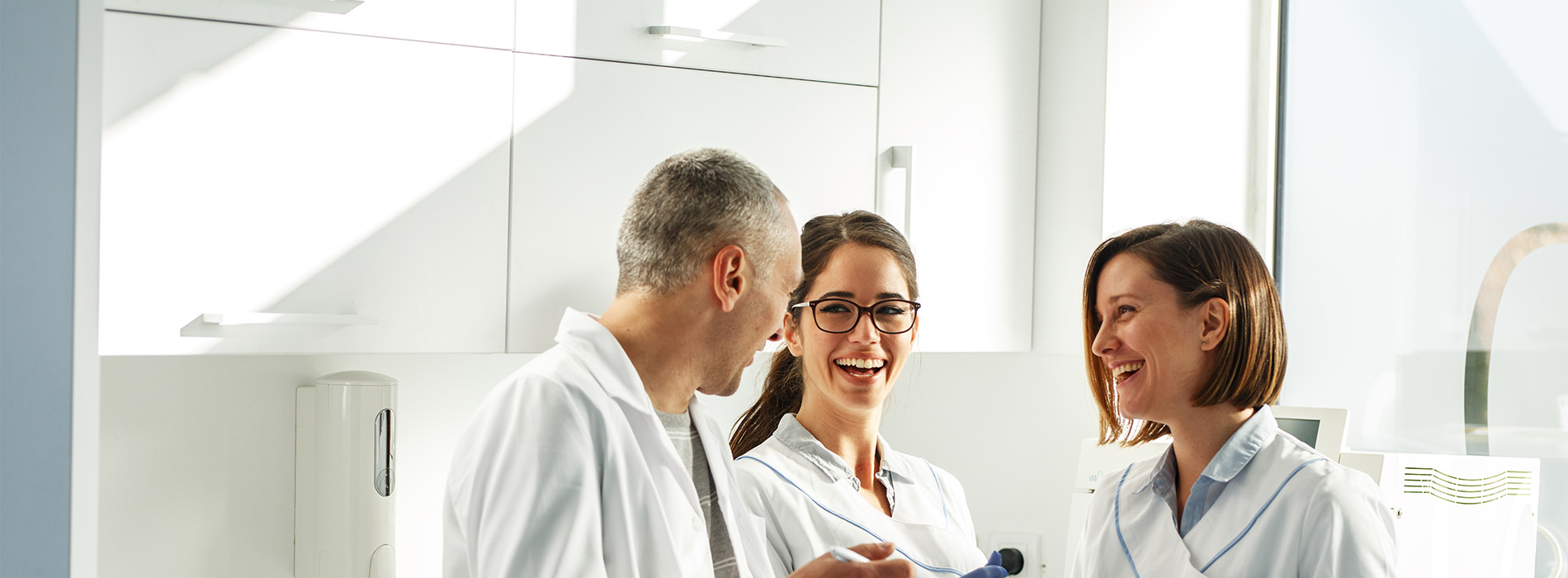 The image shows three individuals in professional attire, likely a medical or healthcare setting, engaging in conversation with smiles on their faces.