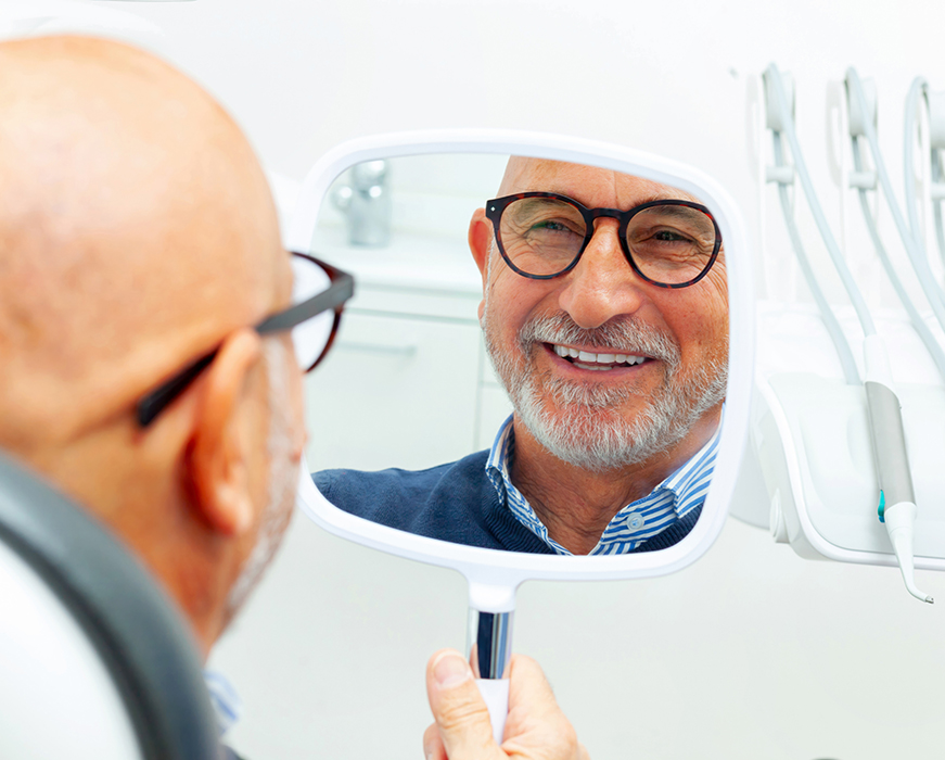 A man with gray hair, wearing glasses and a blue shirt, sits in front of a dental mirror in a dentist s office, smiling at his reflection.