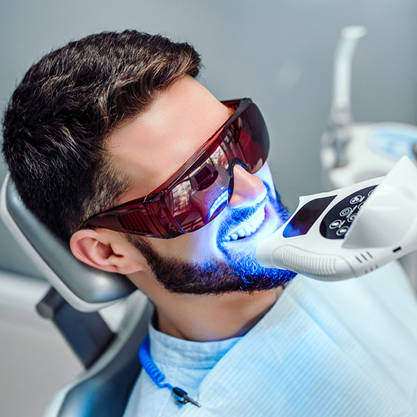 The image shows a man sitting in a dental chair with a bright smile, wearing red-tinted sunglasses, and having his teeth cleaned by a professional using an electronic device.