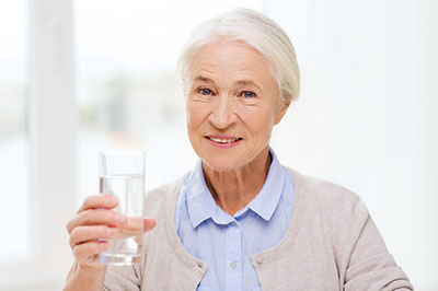 The image depicts an elderly woman holding a clear glass of water, smiling, with her right hand raised slightly above the glass.