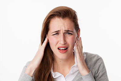 A young woman with blonde hair and a concerned expression holds her hand over her ear, possibly indicating discomfort or hearing issues.
