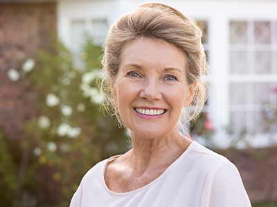 The image shows a woman with light hair smiling at the camera, wearing a white top, standing outdoors in front of a house with a brick facade and a garden area.
