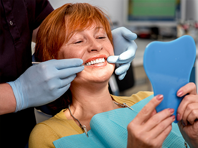 The image shows a woman sitting in a dental chair with a blue mouthguard on her teeth, smiling at the camera while holding up a mirror. A dental professional, wearing gloves and a face mask, is adjusting her teeth with a tool.