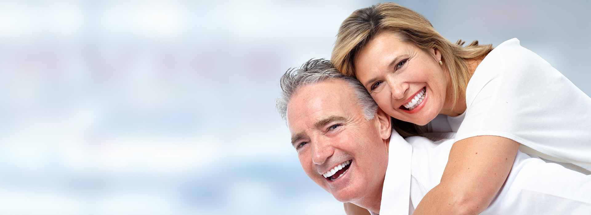 The image shows a man and woman smiling at the camera with their arms around each other, both dressed in white attire.
