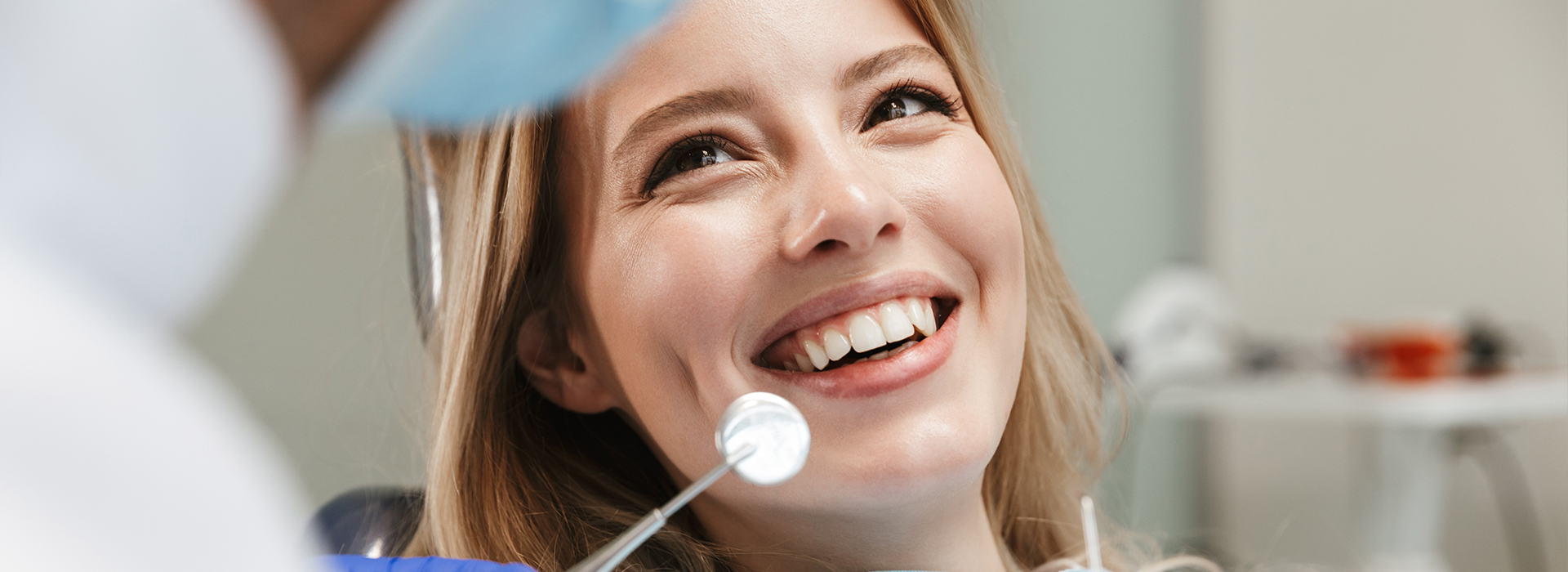 A woman with a bright smile sits in a dental chair, looking directly at the camera, while a dentist examines her teeth.