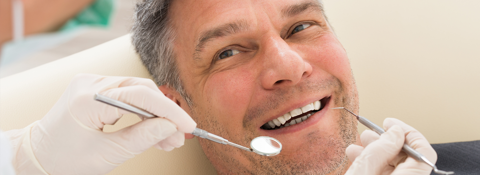 The image shows a man sitting in a dental chair with his mouth open, receiving dental care from a professional, while he smiles and appears relaxed during the procedure.