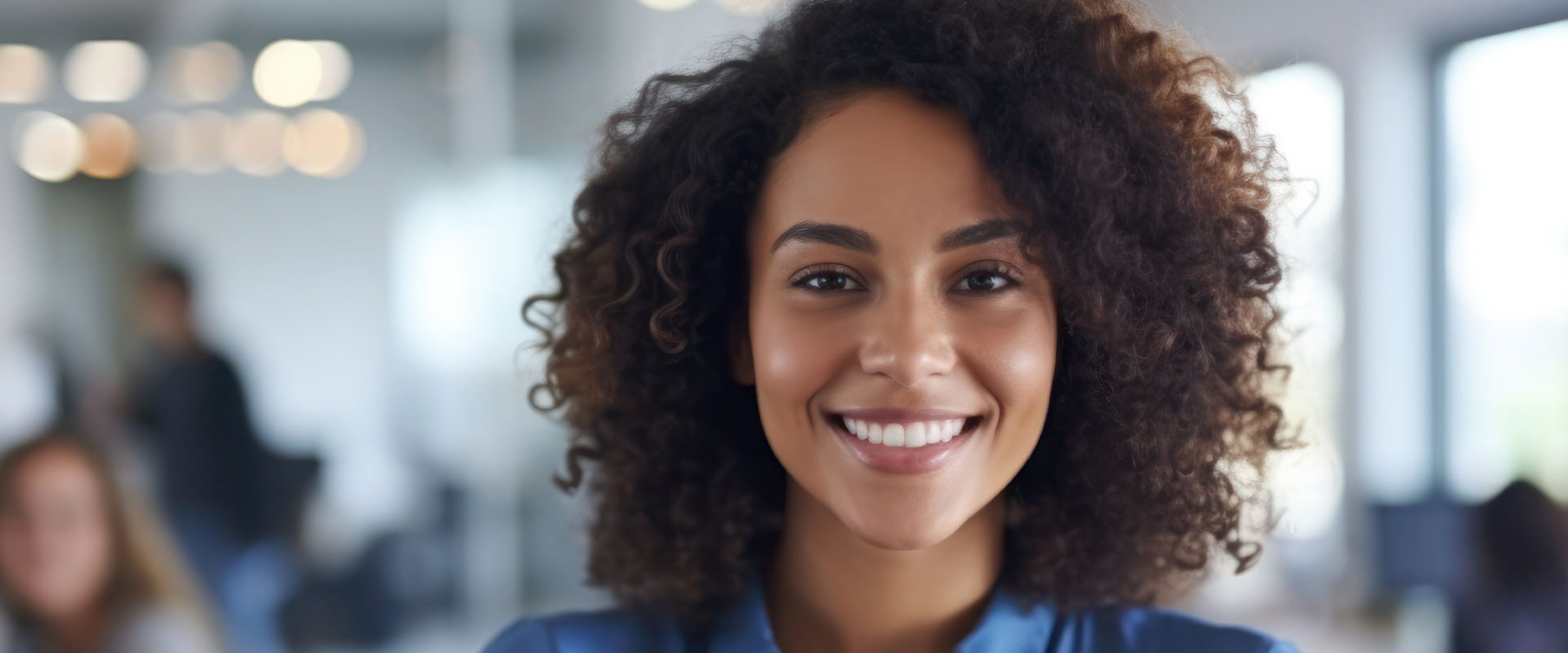 The image shows a person with curly hair smiling at the camera  they are wearing a dark top and have a professional appearance.