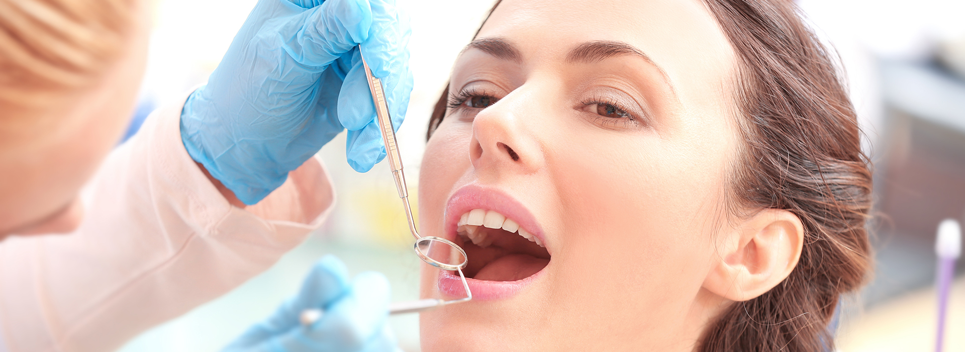 The image shows a woman receiving dental treatment  she is seated with her mouth open while a dentist works on her teeth, using tools visible around her mouth.