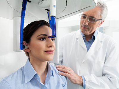 The image shows a woman seated in front of a 3D scanner machine, with a man standing beside her, both in a medical setting.