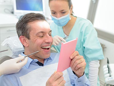 A man sitting in a dental chair, holding up a pink card with a surprised expression, while being attended to by a dental professional.