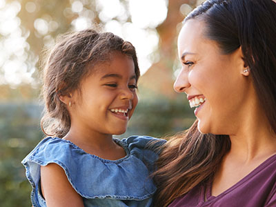 The image shows a woman smiling at her child, who is also smiling, with both standing outdoors during daylight hours.