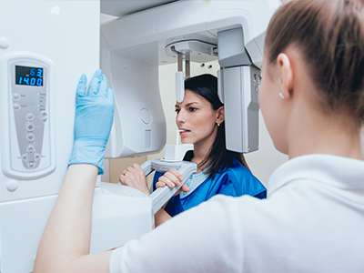 The image shows a woman standing in front of a large 3D scanner with a digital display, while another person stands behind her, observing the scan on the screen. Both individuals are wearing face masks and gloves, suggesting a medical or healthcare setting.