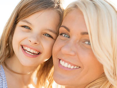 The image shows a woman and a young girl smiling at the camera, with the woman s arm around the child, both appear happy and are outdoors during daylight.