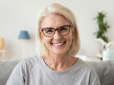 The image shows a woman with blonde hair wearing glasses, smiling at the camera while seated indoors.