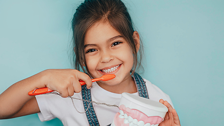 The image shows a young girl brushing her teeth with an electric toothbrush, smiling at the camera.