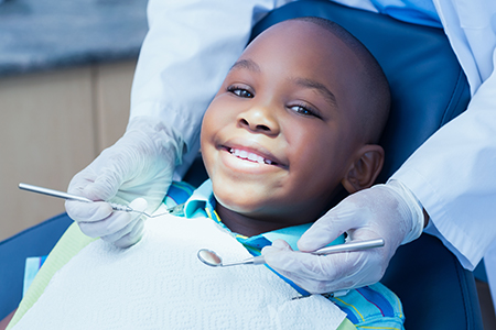 A young boy sitting in a dental chair with a smiling expression, receiving dental care from a professional wearing protective gloves and a face mask.