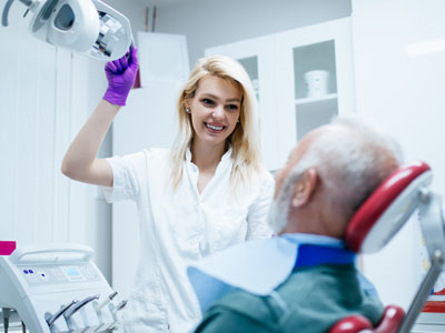A woman in a white coat stands beside a man sitting in a dental chair, holding a dental drill, with both wearing gloves.