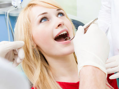 The image shows a woman sitting in a dental chair with her mouth open, receiving dental treatment from a dentist who is performing a procedure on her teeth.