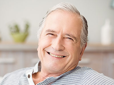 The image shows an older man with white hair, wearing a blue shirt, smiling broadly while sitting comfortably with his hands behind him.
