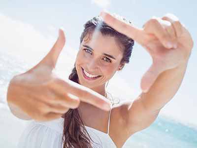 A woman with long hair smiles while holding up her hand to frame the camera lens with her fingers.